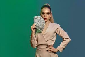 Blonde lady in jewelry and beige dress. She is showing fan of hundred dollar bills, posing on colorful background. Gambling, poker, casino. Close-up photo