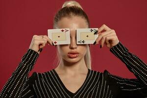 Blonde female in black dress in rhinestones. She closed her eyes with two aces, posing against red background. Gambling, poker, casino. Close-up photo