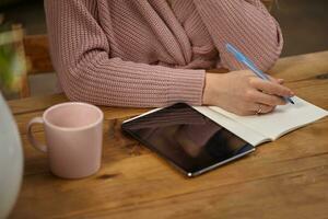 Lady in pink knitted cardigan. She is holding a blue pen, sitting at wooden table with a tablet, notebook and cup on it. Student, blogger. Close-up photo