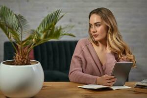 Model blogger in pink cardigan is holding a tablet. Sitting at wooden table with notebooks and palm tree in pot on it. Student, blogger. Close-up photo