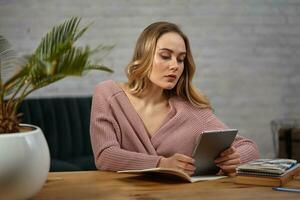 Woman in pink cardigan is holding a tablet. Sitting at wooden table with notebooks, book, blue pen and flower in pot on it. Student, blogger. Close-up photo