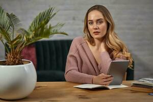 Lady blogger in pink cardigan is holding a tablet. Sitting at wooden table with notebooks and palm tree in pot on it. Student, blogger. Close-up photo