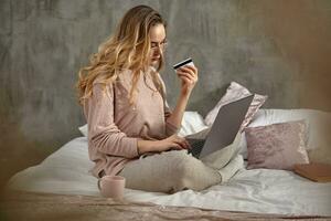 Blonde girl blogger in glasses, casual clothing. Holding plastic card and laptop on her legs, sitting sideways on bed with book and cup. Close up photo