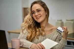 Woman blogger in glasses, beige sweater. Smiling, sitting in kitchen at wooden table, talking by phone, holding notebook and plastic card. Close up photo