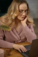 Woman in pink cardigan and glasses. She is propping her face with hand, working on her laptop. Sitting at wooden table with pink cup on it. Close up photo