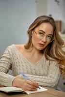 Blonde woman blogger in glasses, beige sweater. Sitting in kitchen at wooden table, going to write something in her notebook, looking at you. Close up photo