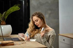 Woman blogger in beige sweater. Sitting in kitchen at table, talking by smartphone, going write something in notebook, holding plastic card. Close up photo