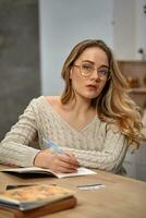 Blonde female blogger in glasses, beige sweater. Sitting in kitchen at wooden table, going to write something in her notebook. Close up photo