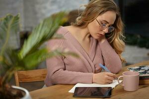 Girl in glasses, pink cardigan. Holding pen, sitting at wooden table with a tablet, notebook, cup and flower in pot on it. Student, blogger. Close-up photo