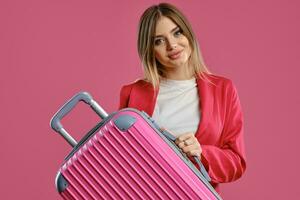 Blonde woman in red pantsuit and white blouse. She smiling, holding suitcase, posing on pink background. Fashion, travelling, advertising. Close-up photo