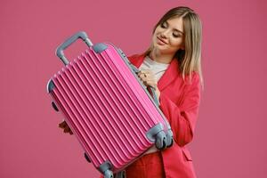 Blonde woman in red pantsuit and white blouse. She smiling, holding suitcase, posing on pink background. Fashion, travelling, advertising. Close-up photo