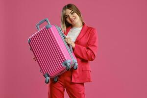 Blonde model in red pantsuit and white blouse. She smiling, holding suitcase, posing on pink background. Fashion, travelling, advertising. Close-up photo