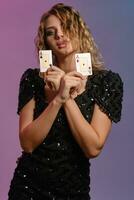 Brown-haired female in black shiny dress showing two playing cards, posing on colorful background. Gambling entertainment, poker, casino. Close-up. photo