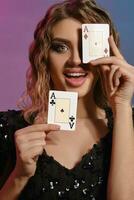 Brown-haired female in black shiny dress is smiling, showing two playing cards, posing on colorful background. Gambling, poker, casino. Close-up. photo