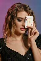 Brown-haired girl in black shiny dress has covered her eye with ace of diamonds, posing on colorful background. Gambling, poker, casino. Close-up. photo