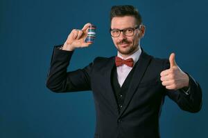 hombre en negro clásico traje, rojo corbata de moño, glases es demostración algunos de colores papas fritas, posando en azul estudio antecedentes. juego, póker, casino. de cerca. foto