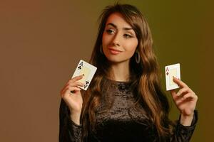Brunette lady in black velvet dress and jewelry is smiling, showing two aces, posing on colorful studio background. Gambling, poker, casino. Close-up. photo