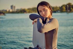 morena niña en un gris suéter tipo con cuello de tortuga tiene cerrado su ojos mientras posando con su wakeboard y en pie en un muelle de el orilla. de cerca. foto