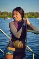 niña en un gris suéter tipo con cuello de tortuga, especial Deportes chaleco y negro reloj es mirando lejos y sonriente mientras posando con su wakeboard en un muelle. de cerca. foto