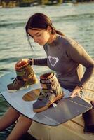 Brunette girl in a gray turtleneck and blue denim shorts is checking up boots on her wakeboard while sitting on a pier of the coastal zone. Close-up. photo