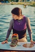 Girl in gray turtleneck, special sports vest is going to go down into the water while posing with her wakeboard on pier of a coastal zone. Close-up. photo