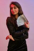 Brunette girl in black dress showing two red chips and some money, posing isolated on white with red backlight. Gambling, poker, casino. Close-up. photo