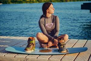 morena niña en un gris suéter tipo con cuello de tortuga, especial Deportes chaleco y azul mezclilla pantalones cortos es posando con su wakeboard en un muelle de el costero zona. de cerca. foto