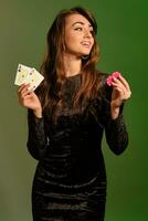 Brunette woman in black dress is smiling, showing two red chips and aces, posing against colorful studio background. Gambling, poker, casino. Close-up photo