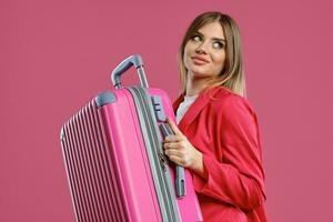 Blonde lady in red pantsuit and white blouse. She smiling, holding suitcase, posing on pink background. Fashion, travelling, advertising. Close-up photo