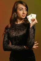 Brunette girl in black velvet dress and jewelry is smiling, showing two aces, posing on colorful studio background. Gambling, poker, casino. Close-up photo