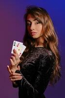 Brunette girl in black velvet dress showing two playing cards, posing sideways on coloful background. Gambling entertainment, poker, casino. Close-up. photo