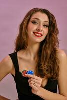 Brunette lady in black dress. She is smiling, showing two colorful chips, posing on pink studio background. Gambling, poker, casino. Close-up photo