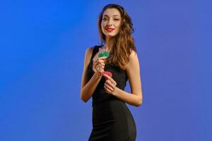 Brunette model with earring in nose, in black dress. She smiling, holding two green and red chips, posing on blue background. Poker, casino. Close-up photo