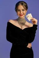 Brunette lady with bare shoulders, in black dress and shiny jewelry. Smiling, showing two aces, posing on purple background. Poker, casino. Close-up photo