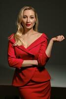 Blonde woman with bright make-up and deep neckline, in red dress and black earrings. She smiling, posing on gray background. Copy space, close up photo