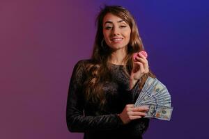 Brunette girl in black velvet dress showing two red chips and some cash, posing against coloful background. Gambling, poker, casino. Close-up. photo