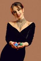 Brunette lady in black dress and brilliant jewelry. Smiling, showing handful of colorful chips, posing on brown background. Poker, casino. Close-up photo