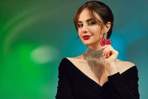 Brunette woman in black dress, necklace and earrings. She is smiling, showing two red chips, posing on colorful background. Poker, casino. Close-up photo