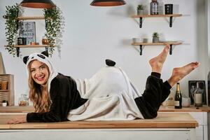 Blonde lady in plush pajama in form of cartoon character panda is having fun at bachelorette party, posing on kitchen table, smiling. Close-up. photo