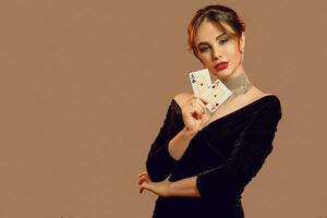 Brunette model, bare shoulders, in black dress and jewelry. Showing two playing cards, posing on brown background. Poker, casino. Close-up photo