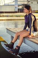 Female in gray turtleneck, special sports vest and denim shorts is posing with a wakebord on her legs sitting on a pier of a coastal zone. Close-up. photo