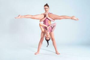 dos flexible muchachas gimnastas con coletas, en rosado leotardos son ejecutando divisiones utilizando apoyo mientras posando aislado en blanco antecedentes. de cerca. foto