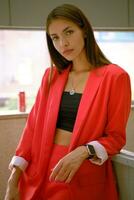 Blonde girl in red lady-type pantsuit and black top, watch, ring, pendant around her neck is posing leaning on a wooden partition. Close-up. photo