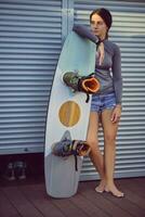 Brunette girl in gray turtleneck and denim shorts is posing with her wakeboard satnding at a coastal zone. Sport and recreation. Close-up. photo