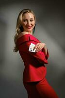 Blonde female in red dress and black earrings. She smiling, showing two aces, posing sideways on gray studio background. Poker, casino. Close-up photo