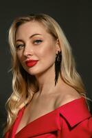 Blonde lady, bright make-up and bare shoulder, in red stylish dress, black earrings. Posing on gray studio background. Close up, copy space photo