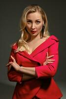 Blonde lady with bright make-up and deep neckline, crossed hands, in red dress and black earrings. She is posing against gray background. Close up photo