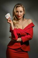 Blonde lady in red dress and black earrings. She smiling, showing two playing cards, posing on gray studio background. Poker, casino. Close-up photo