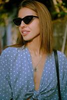 Blonde girl in long blue dress, sunglasses and a small black handbag on her shoulder is walking alone in the city. Fashion and style. Close-up shot. photo