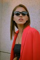 Blonde girl in a red lady-type pantsuit and black top, pendant and sunglasses is posing near a stone wall. Fashion and style. Close-up. photo